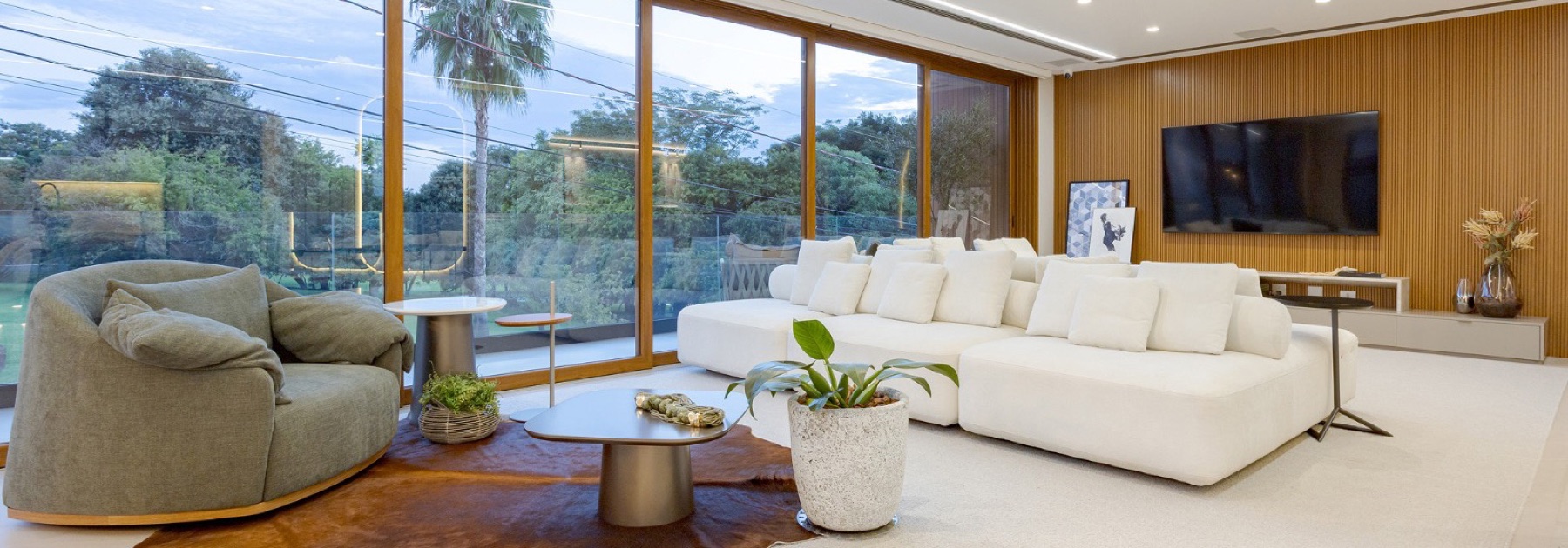 Sala de estar com sofá modular branco, poltrona e grandes janelas de vidro com vista para o jardim